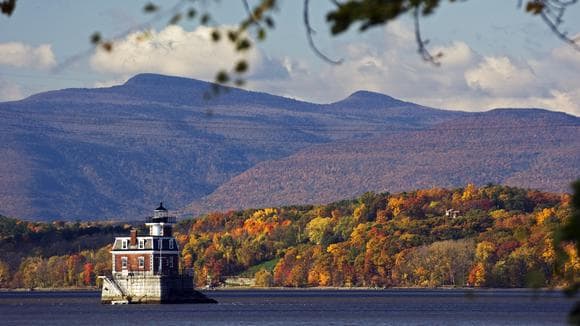 A historic lighthouse is positioned on the water, surrounded by vibrant autumn foliage. Soft mountains rise in the background, creating a serene landscape under a partially cloudy sky.