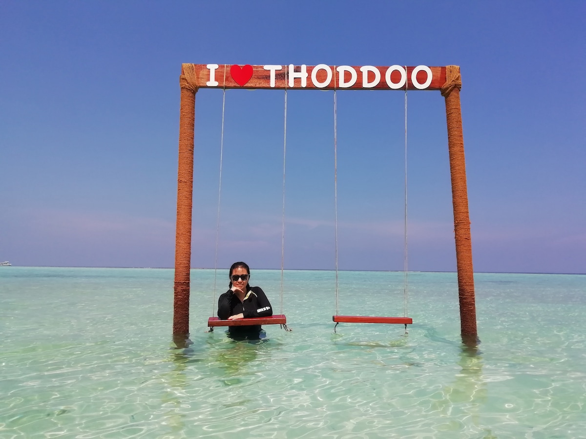 A wooden swing with the text 'I ❤️ Thoddoo' is positioned in shallow turquoise waters, surrounded by gentle waves. A person is standing in the water, holding onto the swing. The clear blue sky complements the serene beach setting.