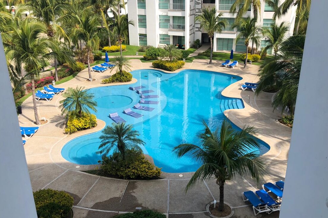A spacious swimming pool is surrounded by palm trees, with several lounge chairs arranged along the perimeter. The pool features a unique shape, and pathways create a clear layout. Lush green landscaping enhances the serene environment of the residential area.