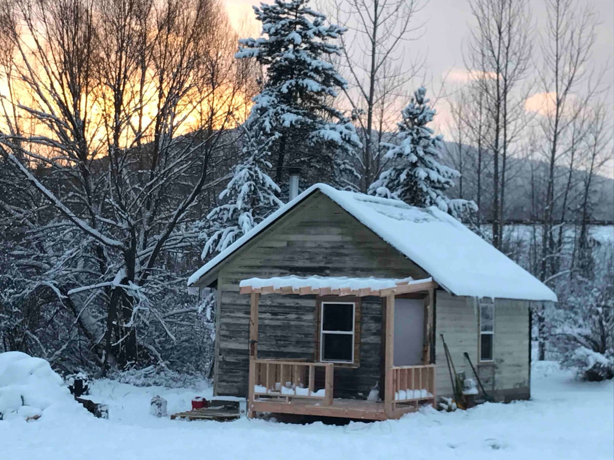 A rustic cabin is framed by snow-covered trees and a wintry landscape. The structure features a small porch with wooden railing, and a window reflects the warm hues of a sunset in the background. Snow blankets the ground, creating a serene winter scene.
