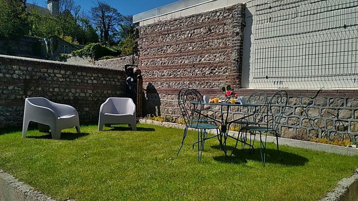 A shared garden area features a well-maintained lawn, surrounded by stone walls. A small round dining table is accompanied by four chairs, while two modern lounge chairs are positioned nearby for relaxation. Colorful flowers add a touch of vibrancy to the space.