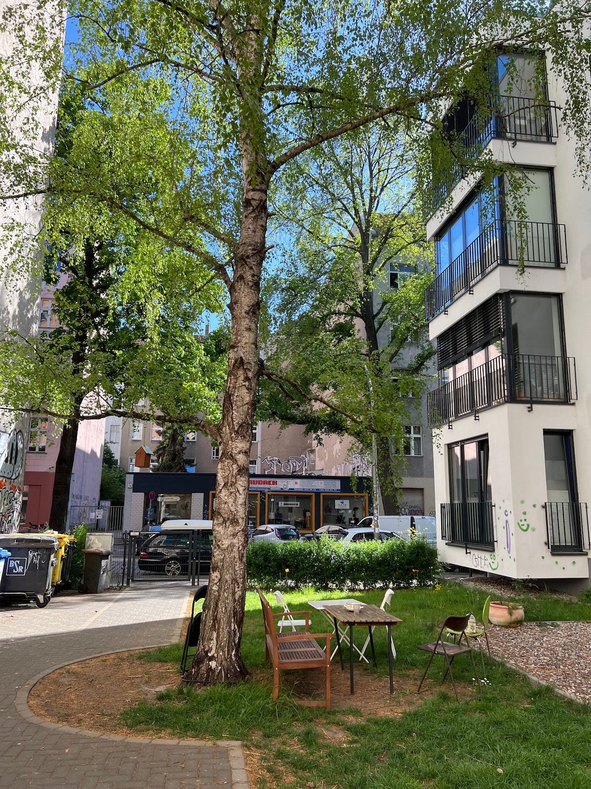 A serene outdoor garden space is highlighted by a large, leafy tree providing shade. Two wooden chairs and a small table are placed on the grass, creating a cozy spot for relaxation. The surrounding greenery offers a calm atmosphere amid the urban setting.