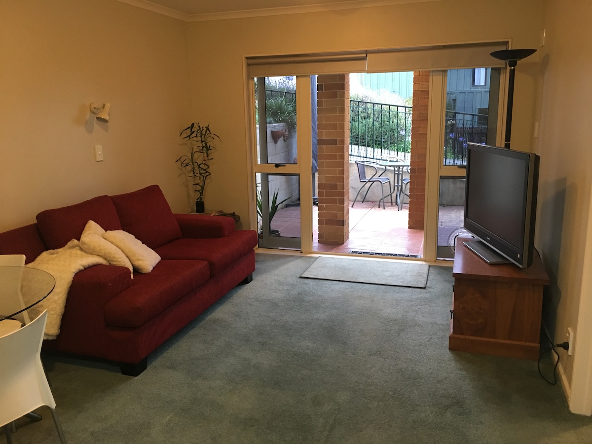 A welcoming living area features a red sofa with decorative pillows facing a flat-screen television. A glass dining table sits beside the sofa, and large sliding doors lead to a private patio. Natural light filters into the space, highlighting the soft blue carpet.