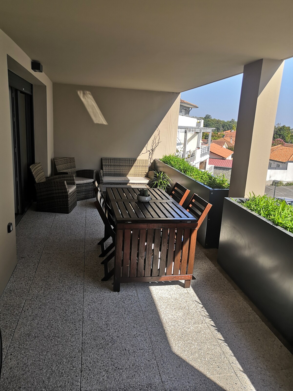 A spacious terrace is shown, featuring a wooden dining table accompanied by four chairs. Two comfortable seating arrangements are visible in the corner. Lush greenery lines the edge, and the scene is well-lit by natural sunlight filtering in.