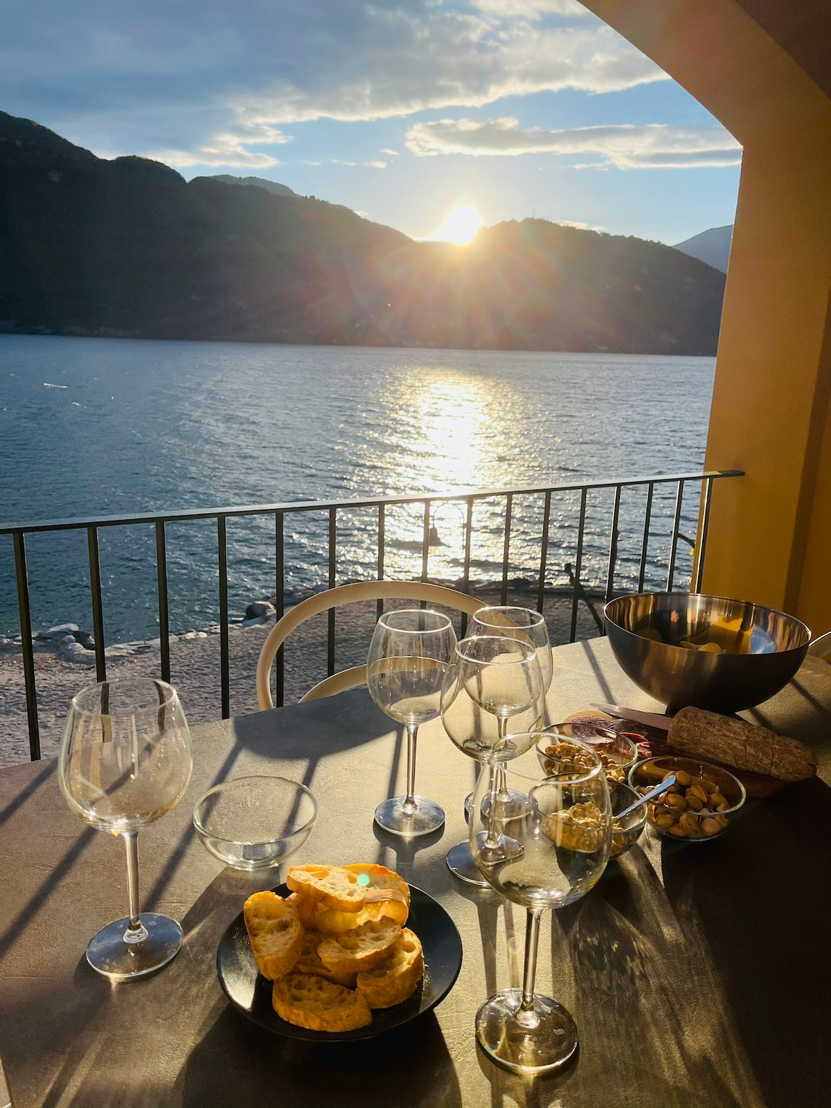 A table set for enjoyment overlooks a sparkling lake, with sunlight reflecting off the water. Glasses are arranged alongside a bowl of snacks and a plate of pastries, framed by the arched balcony. Mountains rise in the background, creating a serene setting.