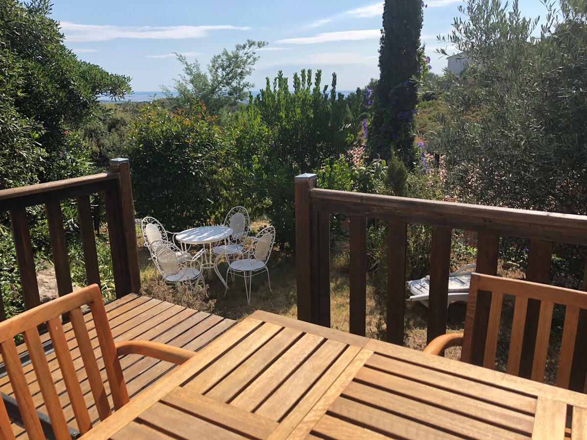 A private terrace features a wooden table surrounded by four chairs, offering a view of a small garden with lush greenery. In the distance, a glimpse of the sea can be seen through the trees, providing a serene outdoor space.