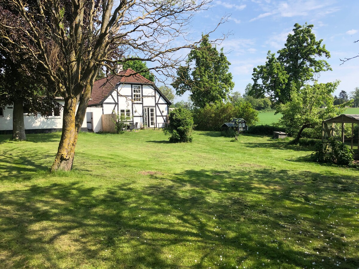 A charming country house is set on a spacious lawn, surrounded by trees and greenery. The building's traditional architecture is visible, complemented by a serene backdrop of fields and a clear blue sky. Outdoor seating areas can be seen among the landscape.
