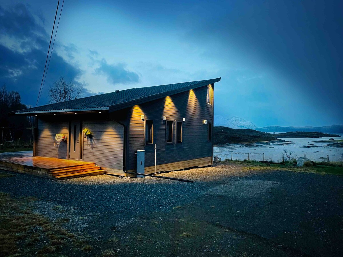 The exterior of the cabin is shown during twilight, with soft lighting illuminating its modern design. A wooden deck leads to the entrance, while the surrounding landscape features rocky ground and views of water and distant mountains.