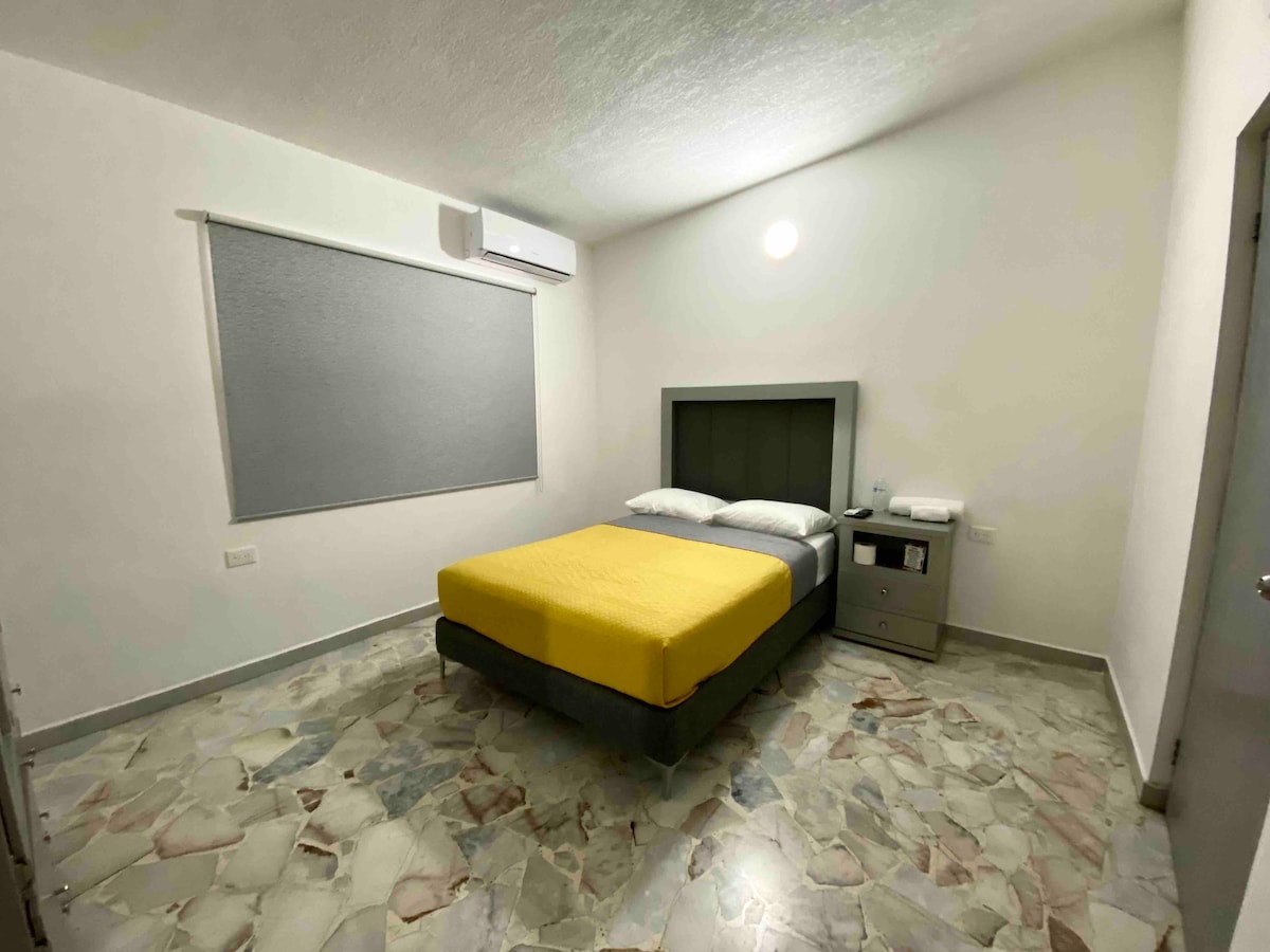 A bright and minimalistic room features a spacious bed with yellow bedding and a dark headboard. A nightstand is positioned beside the bed, while a window with a gray blind allows natural light into the space. The floor is covered in light-colored tiles.
