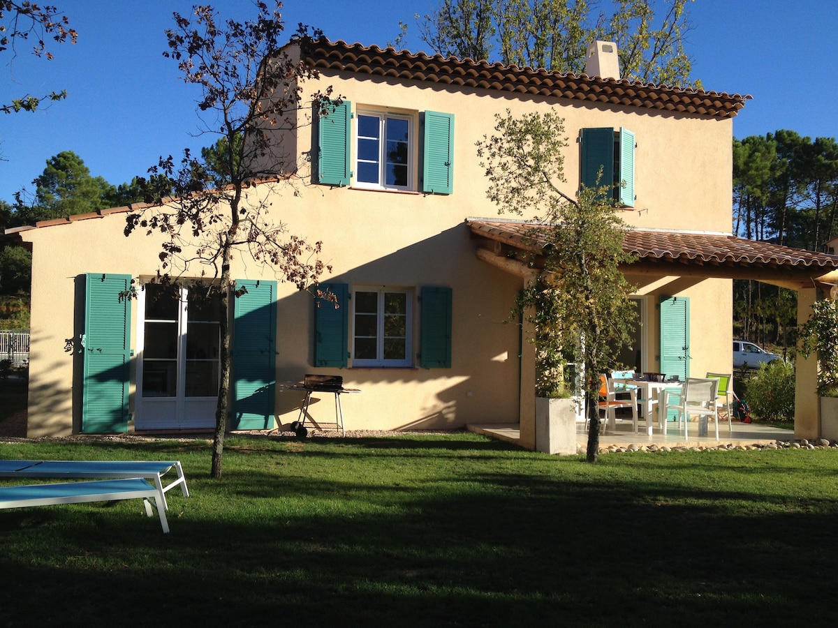 A two-story villa is depicted, showcasing light-colored walls and turquoise shutters. A covered terrace is visible, furnished with a dining table and chairs, while a barbecue grill is present. Lush green grass surrounds the property, with sunlight streaming down, enhancing the vibrant outdoor space.