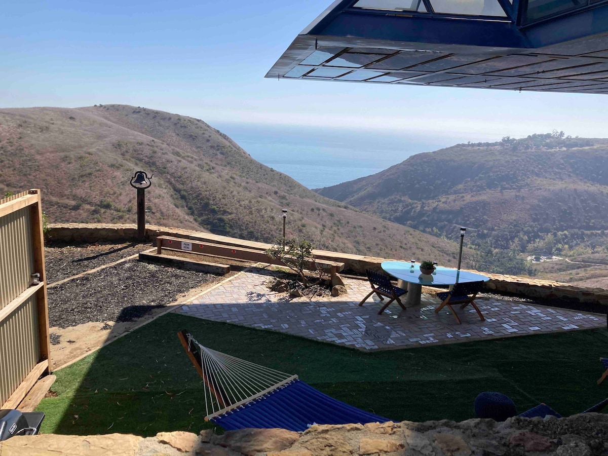 The outdoor seating area features a round table surrounded by two chairs, set on a patio overlooking rolling hills and the ocean. A hammock is seen in the foreground, and lush greenery complements the scenic landscape.