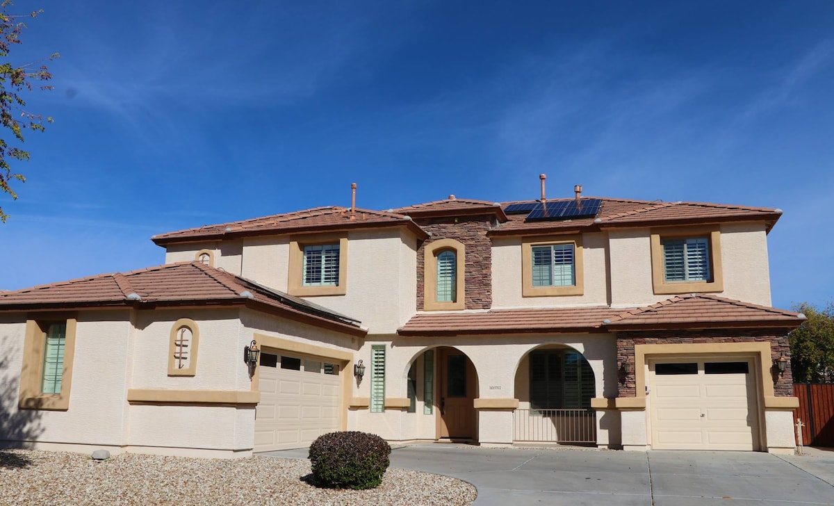 The two-story home features a stucco exterior with a combination of light and dark tones. A driveway leads to two garage doors, while decorative stone accents add character. Bright blue skies frame the house, highlighting large windows and a landscaped area with gravel and a shrub.