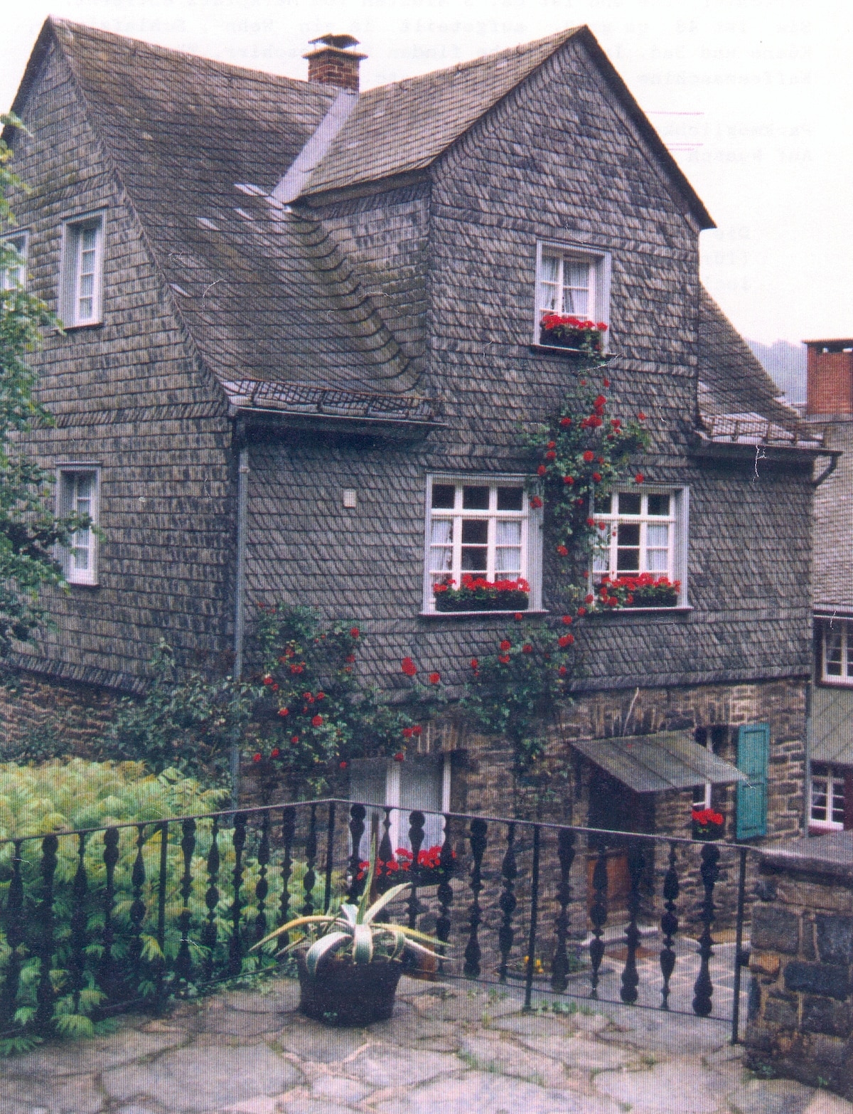 A historic building features a distinctive slate roof and is adorned with red flowers in window boxes. The structure is set against a lush green backdrop, and a stone terrace with a wrought-iron railing provides an inviting outdoor space.