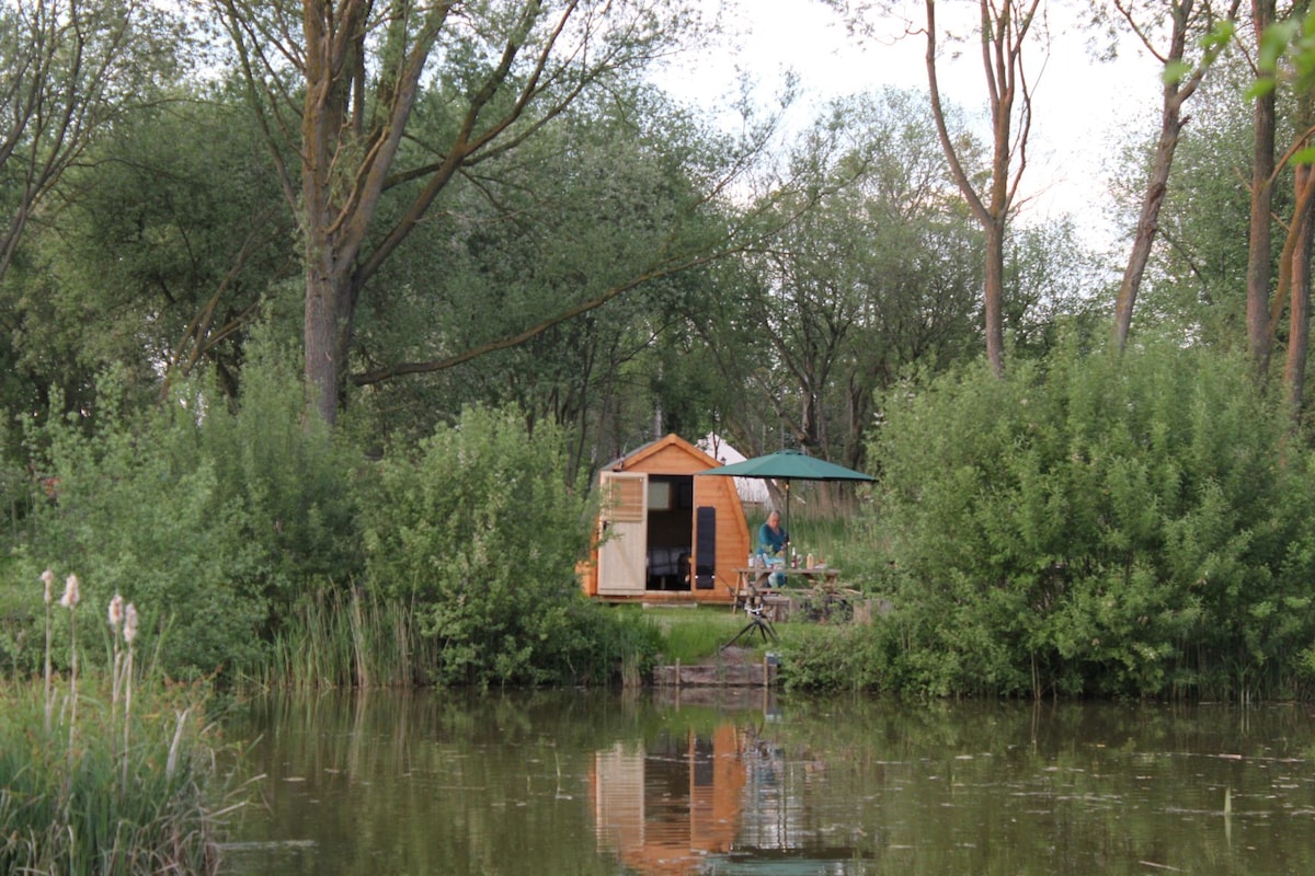 A charming lakeside pod is nestled among lush greenery, with a shaded seating area by the water's edge. The reflection of the pod and surrounding trees can be seen in the calm lake, enhancing the peaceful atmosphere of the retreat.