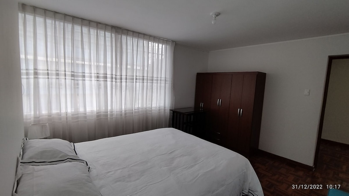 A serene bedroom features a spacious bed with white linens, accompanied by a bedside lamp. Natural light fills the room through large, sheer curtains. A wooden wardrobe and a dark dresser provide ample storage, enhancing the room's practical layout.