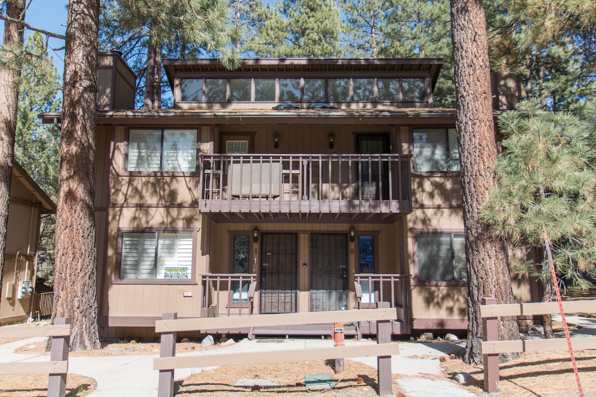 A three-story condominium is nestled among tall pine trees, featuring a balcony on the upper level. The exterior is clad in inviting brown wood, complemented by large windows that flood the interior with natural light.