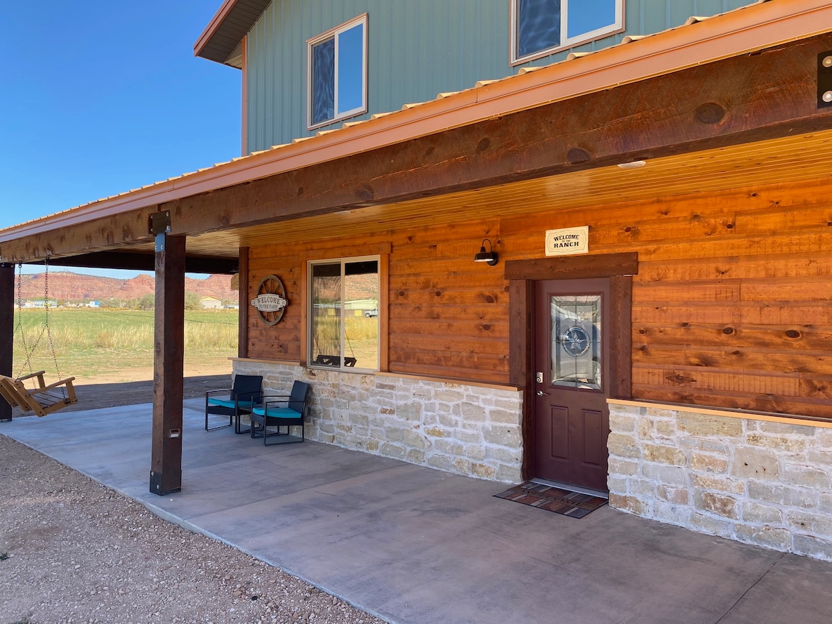 The entrance is marked by a wooden door with a decorative glass insert, surrounded by natural wood paneling. A covered porch features comfortable seating and adjacent porch swings, with expansive views of the surrounding fields and distant red cliffs under a clear blue sky.