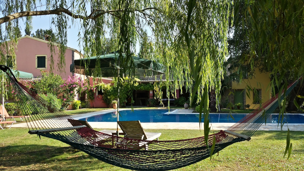 A hammock is suspended between trees, overlooking a private pool surrounded by colorful foliage and blooming flowers. Sun loungers are positioned nearby on a grassy area. The two houses can be seen in the background, offering a glimpse of the villa's inviting outdoor space.