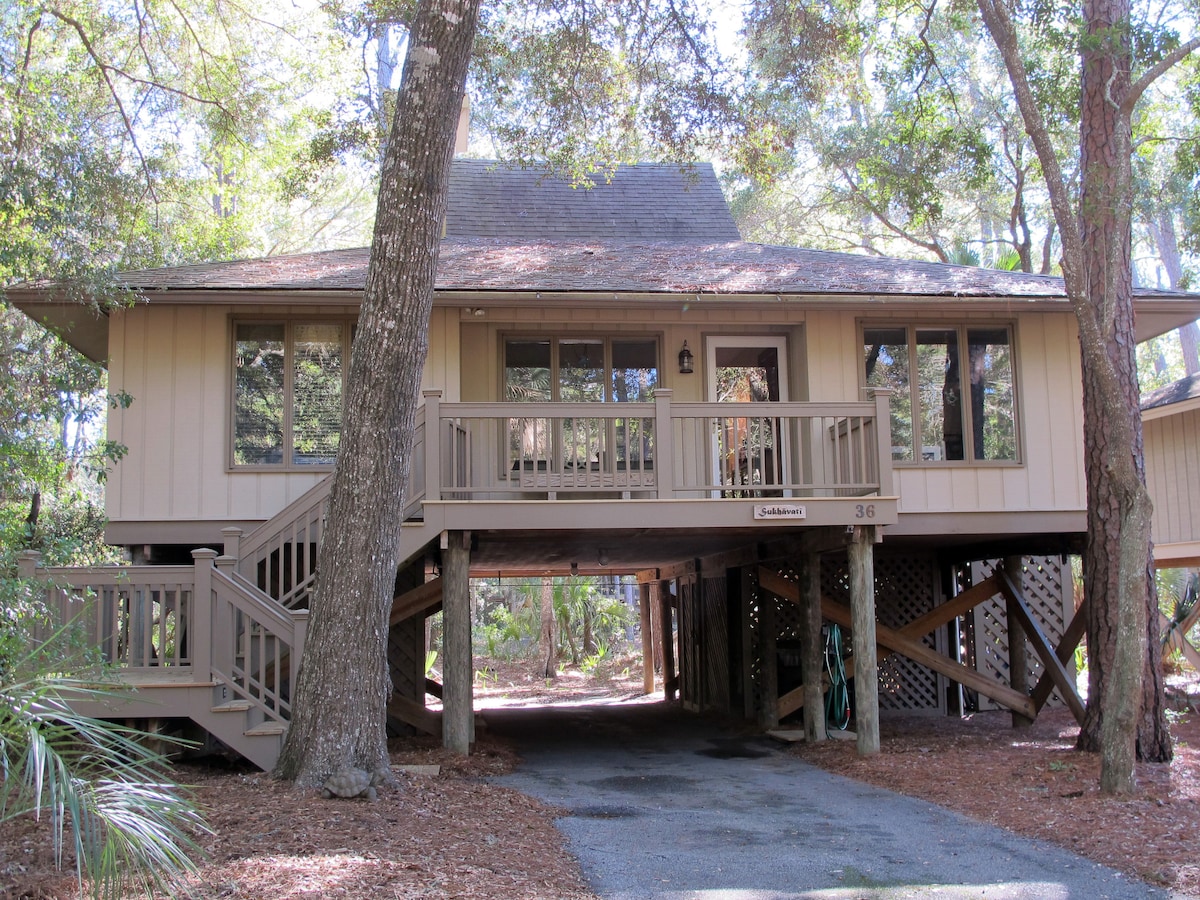 A cottage is elevated on wooden stilts, surrounded by lush trees and natural landscaping. A welcoming front porch with railings is visible, along with large windows that allow for natural light. The entrance features a small light fixture and is easily accessible by an adjacent pathway.