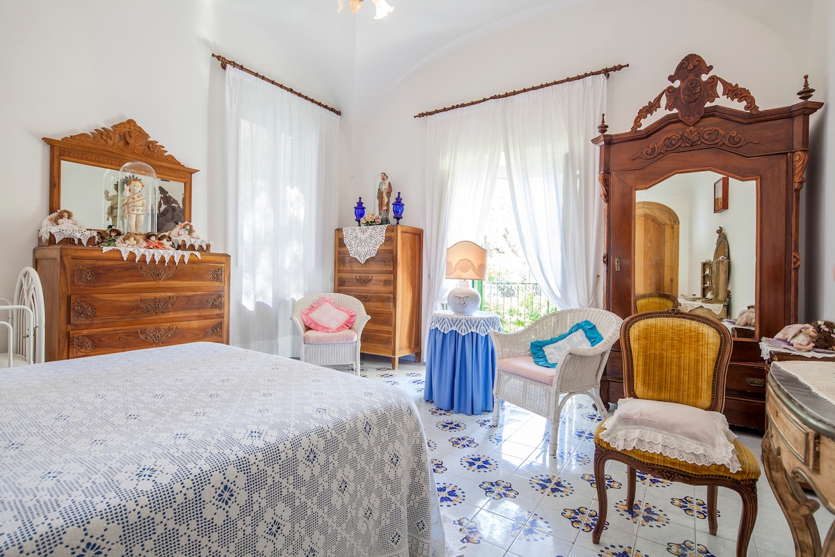 A bright bedroom features a spacious bed adorned with a delicate lace coverlet. Antique wooden furniture includes a large mirror, a dresser, and a vintage chair, complemented by a small table with a blue cloth. Natural light filters through the curtains, creating a serene atmosphere.