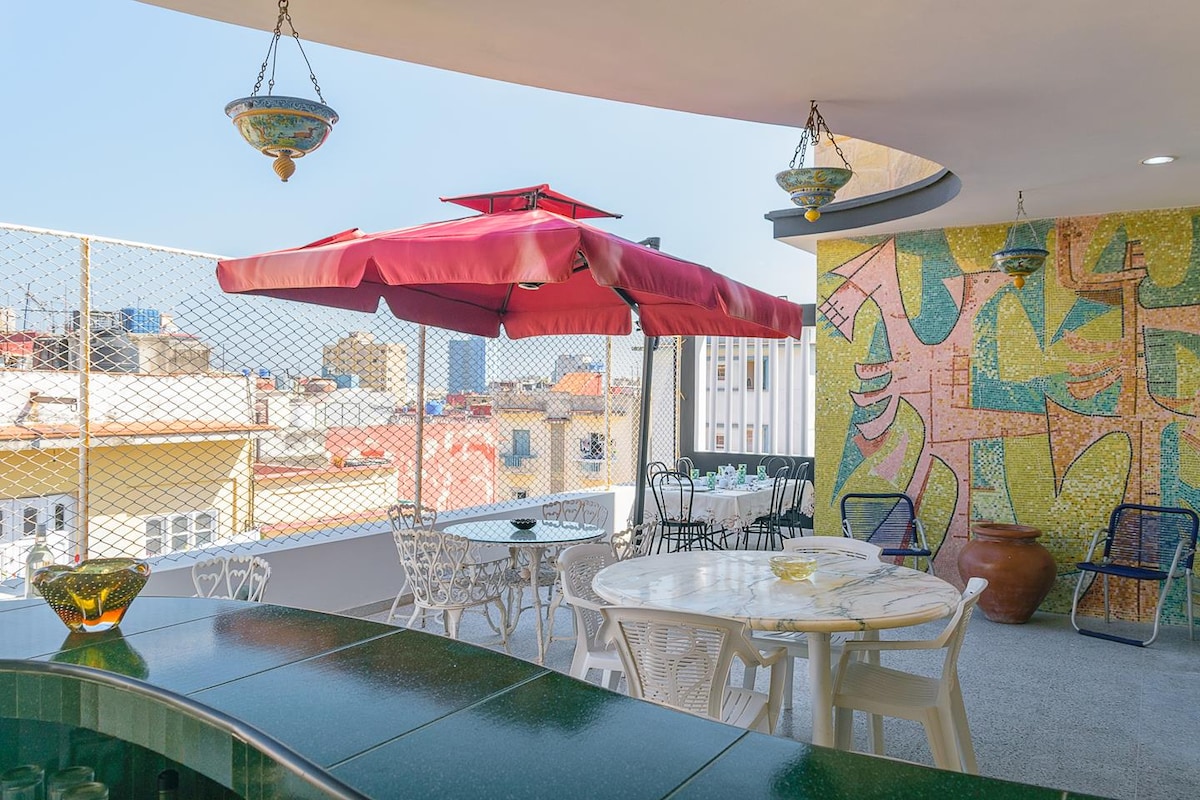 A terrace showcases a vibrant mural adorned with abstract shapes. Seating is arranged around a round table, accompanied by white metal chairs. A large umbrella provides shade, while decorative hanging lights enhance the open space with a view of the city.