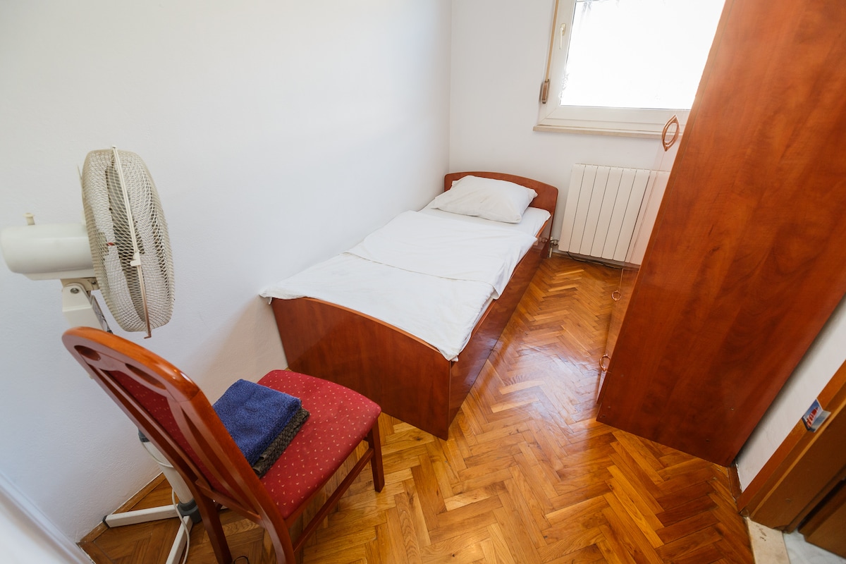 A single bed is positioned against a white wall, accompanied by neatly arranged towels on a nearby chair. A ceiling fan offers comfort, while natural light filters through a window, enhancing the clean and simple design of the room.