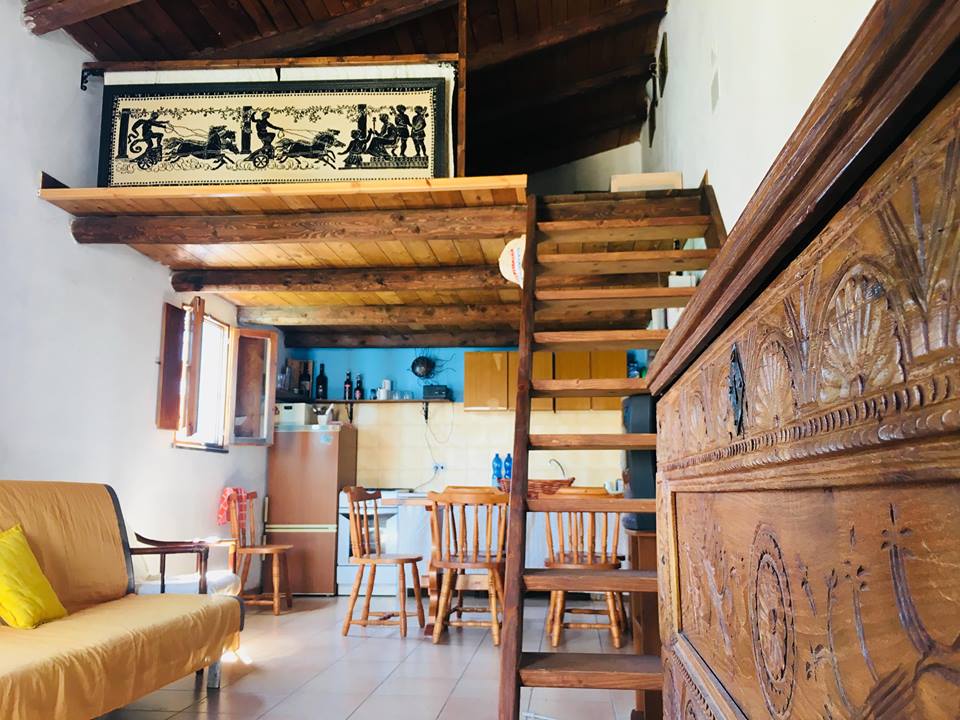 An open living area features a wooden staircase leading to a loft space. Rustic wooden beams extend across the ceiling, while a hand-carved wooden chest and a sofa are positioned on the side. A dining table with chairs sits adjacent to a small kitchen area.