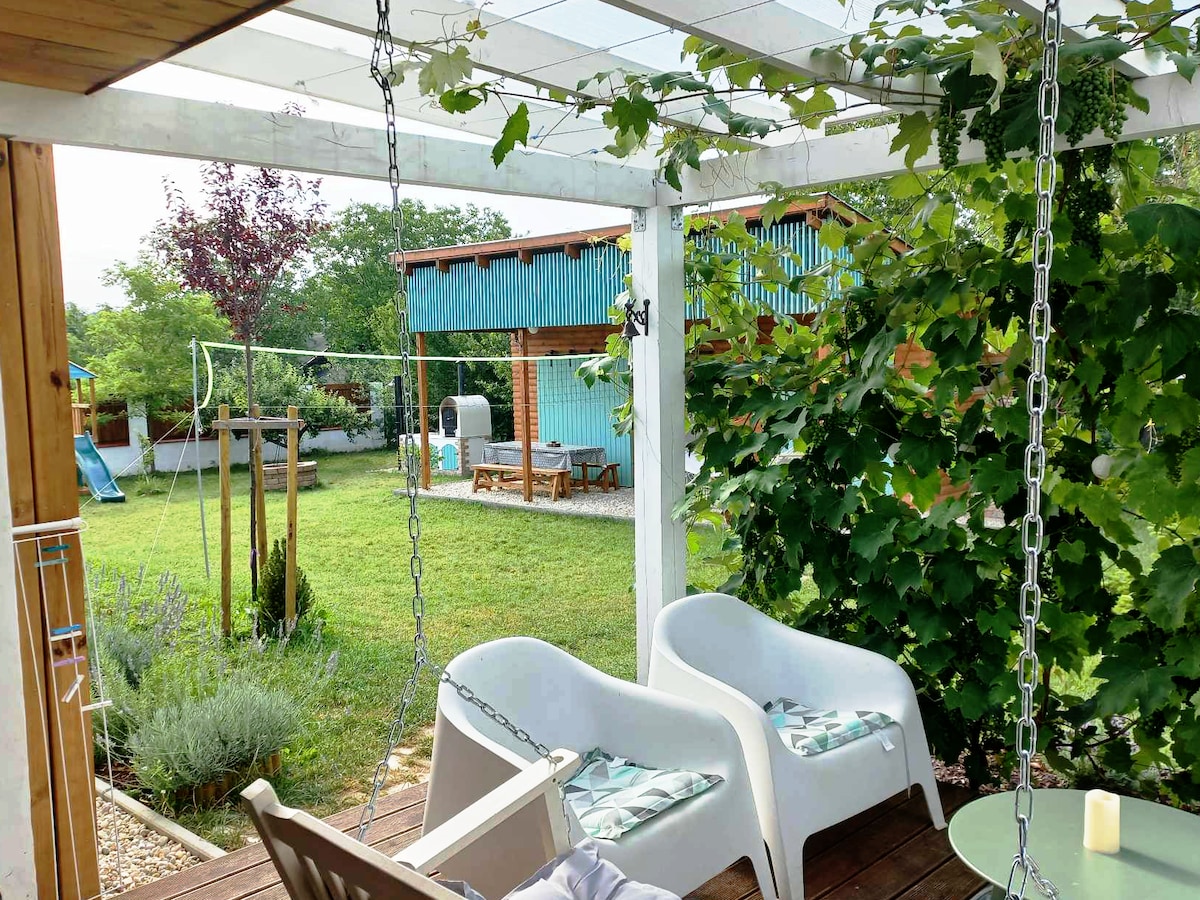 A covered terrace offers two white, hanging seats paired with a small table. Lush greenery surrounds the area, while a spacious lawn is visible in the background, featuring seating arrangements and recreational elements.