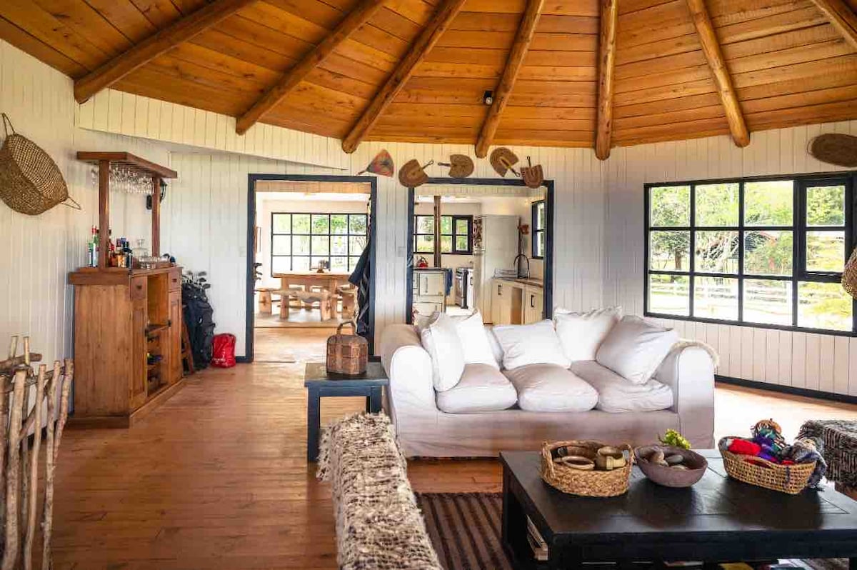 A spacious circular living area showcases a wooden ceiling with exposed beams. A large, light-colored sofa is positioned in front of a dark coffee table adorned with woven baskets. Views of the exterior are provided by multiple large windows, contributing to a bright and airy ambiance.