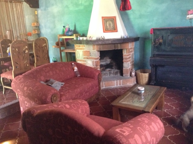 A cozy living area features a traditional fireplace with a stone surround and a wooden beam overhead. Two red upholstered sofas are positioned for comfort, and a small wooden coffee table rests in the center. A piano and a dining area can be seen in the background.