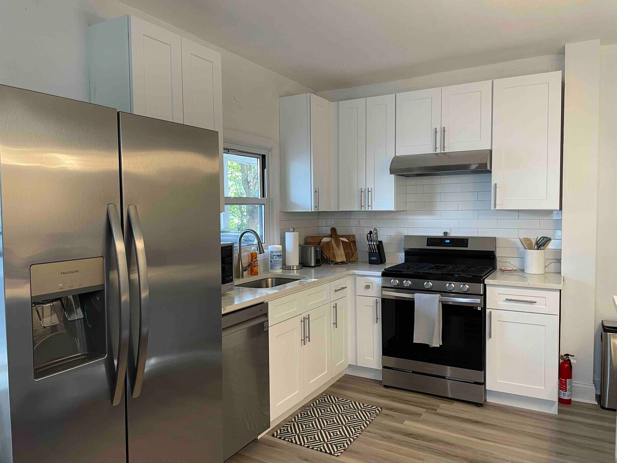 The kitchen features modern stainless steel appliances, including a large refrigerator and a gas stove. White cabinetry complements the bright countertops, while a tiled backsplash adds a clean finish. Natural light filters in through a window above the sink, enhancing the open layout.