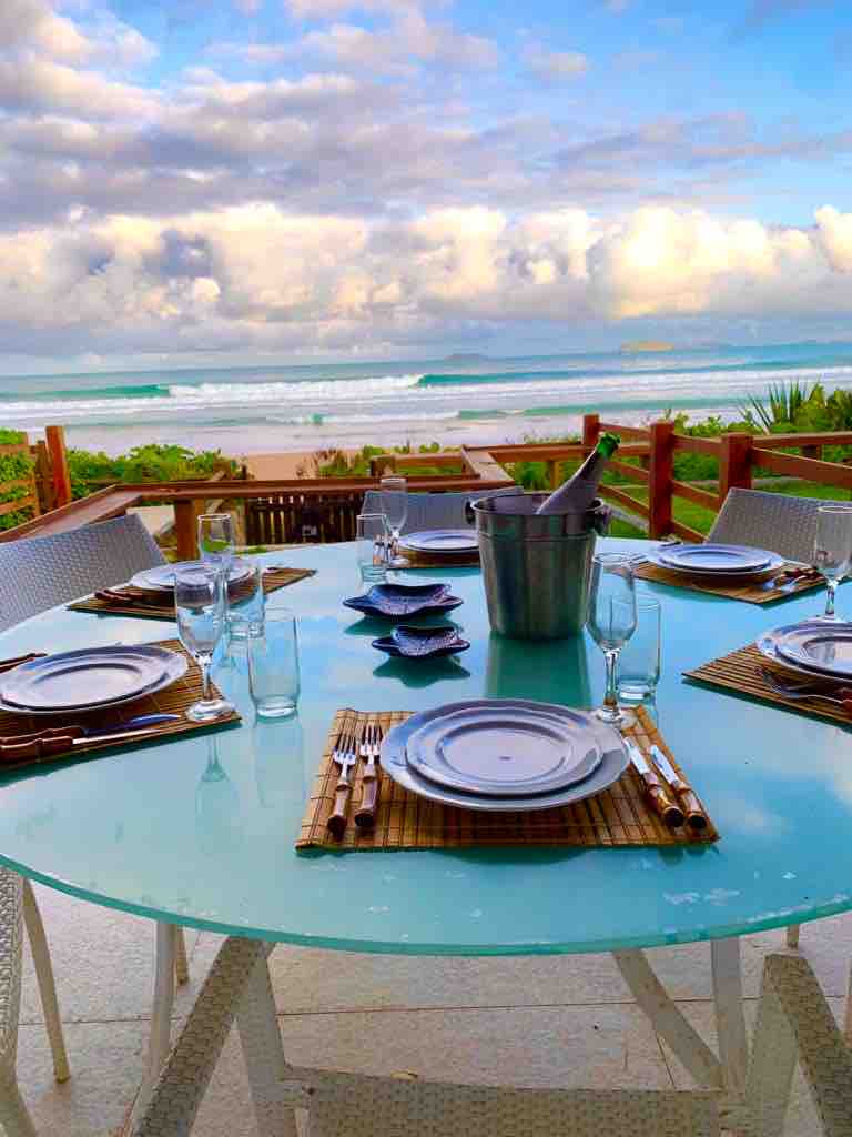 A round dining table is set for a meal with elegant glassware and plates arranged neatly. A cooler sits in the center, with ocean waves visible in the background, creating a serene coastal atmosphere. Lush greenery frames the view, enhancing the outdoor dining experience.