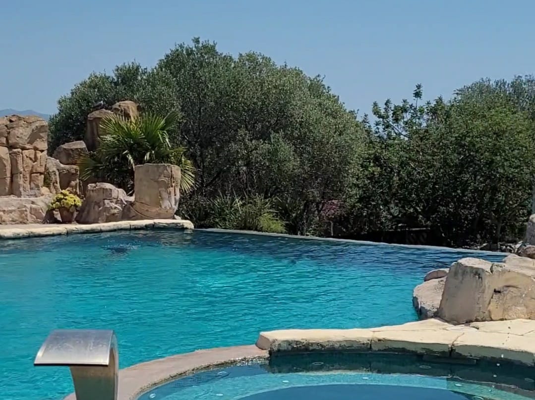 A tranquil outdoor pool is surrounded by lush greenery and rocky landscaping. The clear blue water reflects the sunlight, inviting relaxation. A spa area with a circular design is visible on the edge of the pool, enhancing the serene outdoor environment.