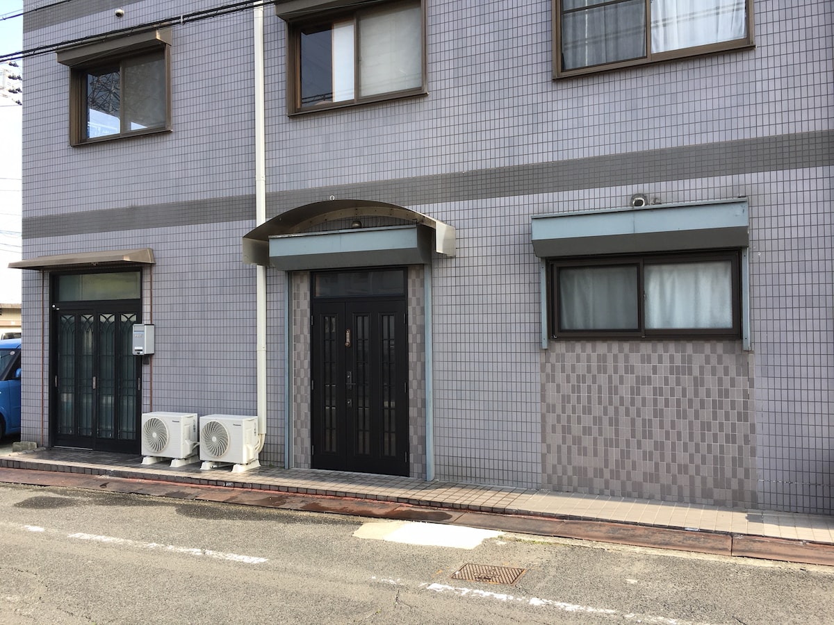 The exterior of the building features a simple facade with a mix of light and dark tiles. A black entrance door is flanked by large windows, and there are two air conditioning units visible on the ground level. The entrance is set back slightly from the street.