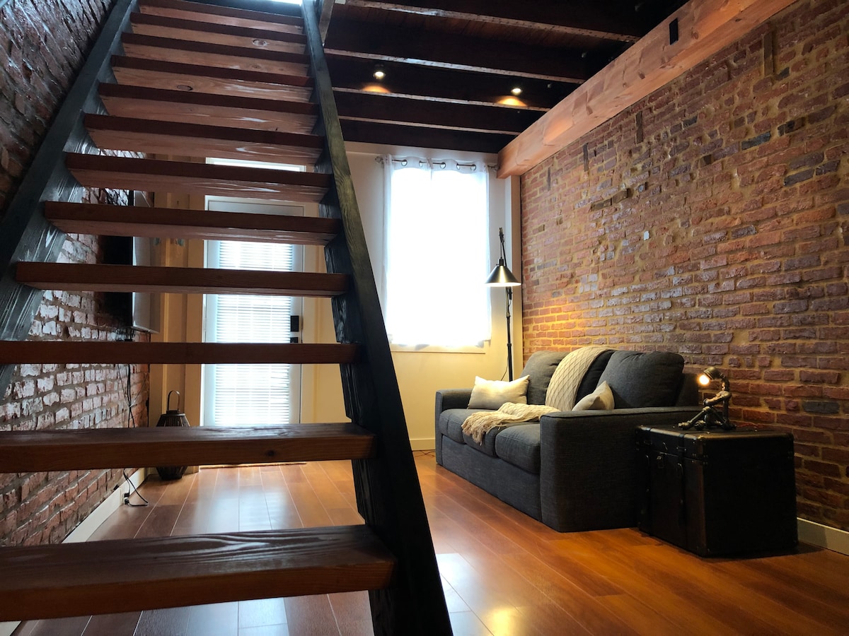 The living space features exposed brick walls that add character, complemented by wooden beams across the ceiling. A comfortable gray sofa is positioned next to a small table and stylish lamp. Wide wooden steps lead to a higher level, enhancing the open layout.