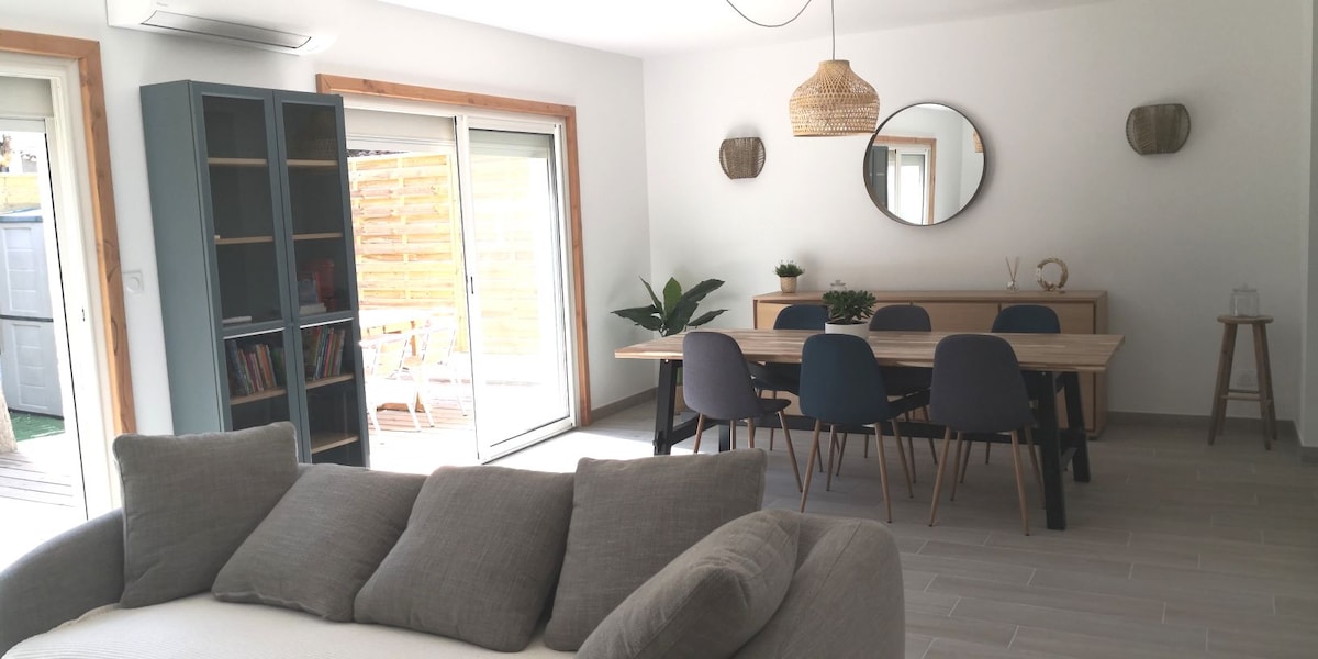 An open living space is presented, featuring a light-colored sofa in the foreground. A dining table with several chairs occupies the center, while a bookshelf is situated against a wall. Large glass doors allow natural light and provide access to the outdoor area.