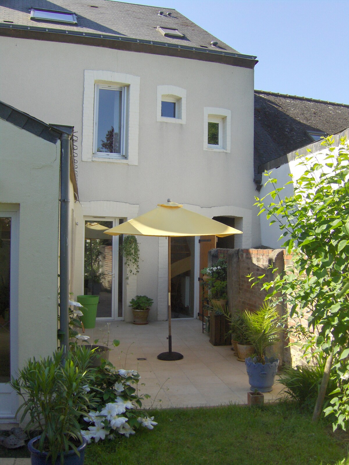 A serene garden terrace is featured, highlighted by a yellow umbrella providing shade. The area is surrounded by various potted plants, including green foliage and white lilies, against a backdrop of the house's light-colored exterior.