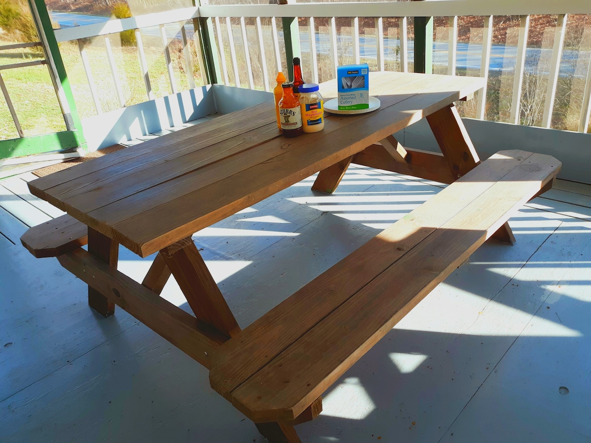 A wooden picnic table is positioned on a spacious screened-in porch, surrounded by natural light. Condiments and beverages are arranged on the table, creating a welcoming space for outdoor dining. The floor features light-colored wood planks, adding to the rustic charm.