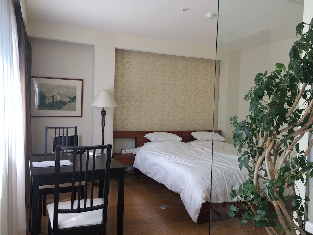A well-appointed room features a spacious bed with crisp white bedding, complemented by a dining area with a black table and chairs. A decorative plant adds a touch of greenery, while large windows allow natural light to fill the space.