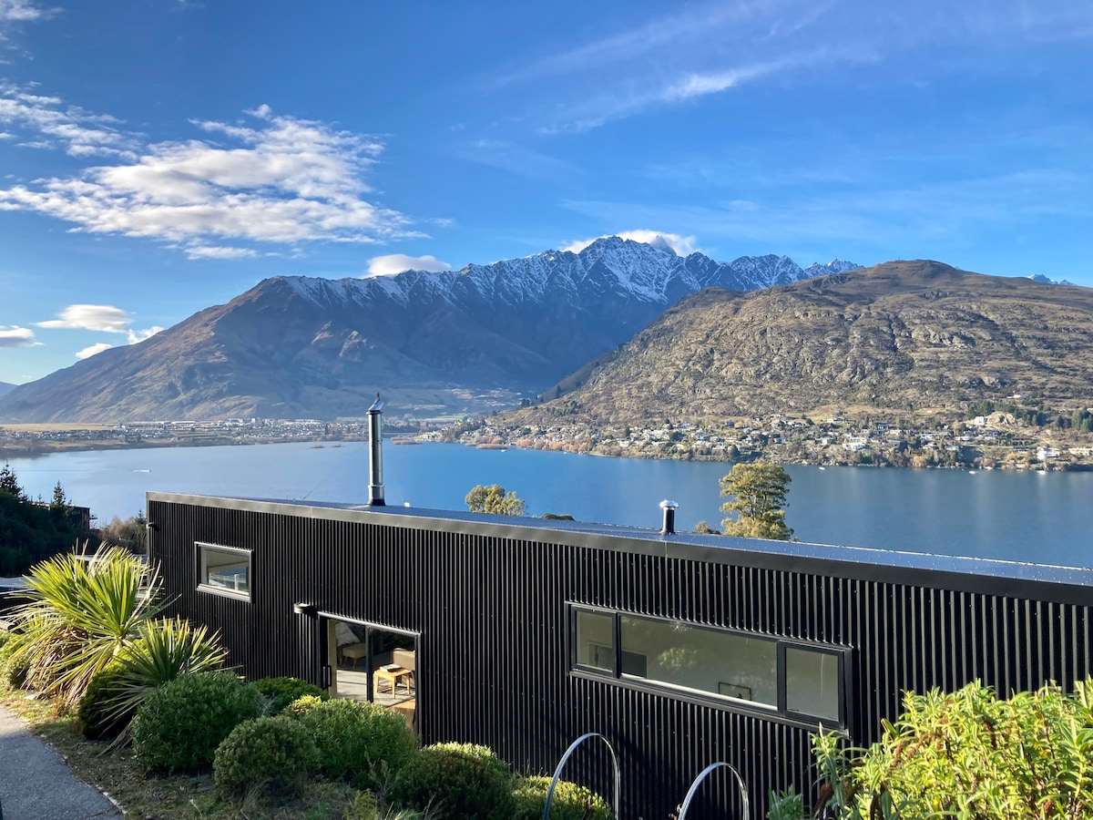 A modern house with a black exterior is positioned on a hillside overlooking Lake Wakatipu. Snow-capped mountains rise in the background, while the serene lake reflects the sky. Lush greenery surrounds the property, contributing to its tranquil setting.