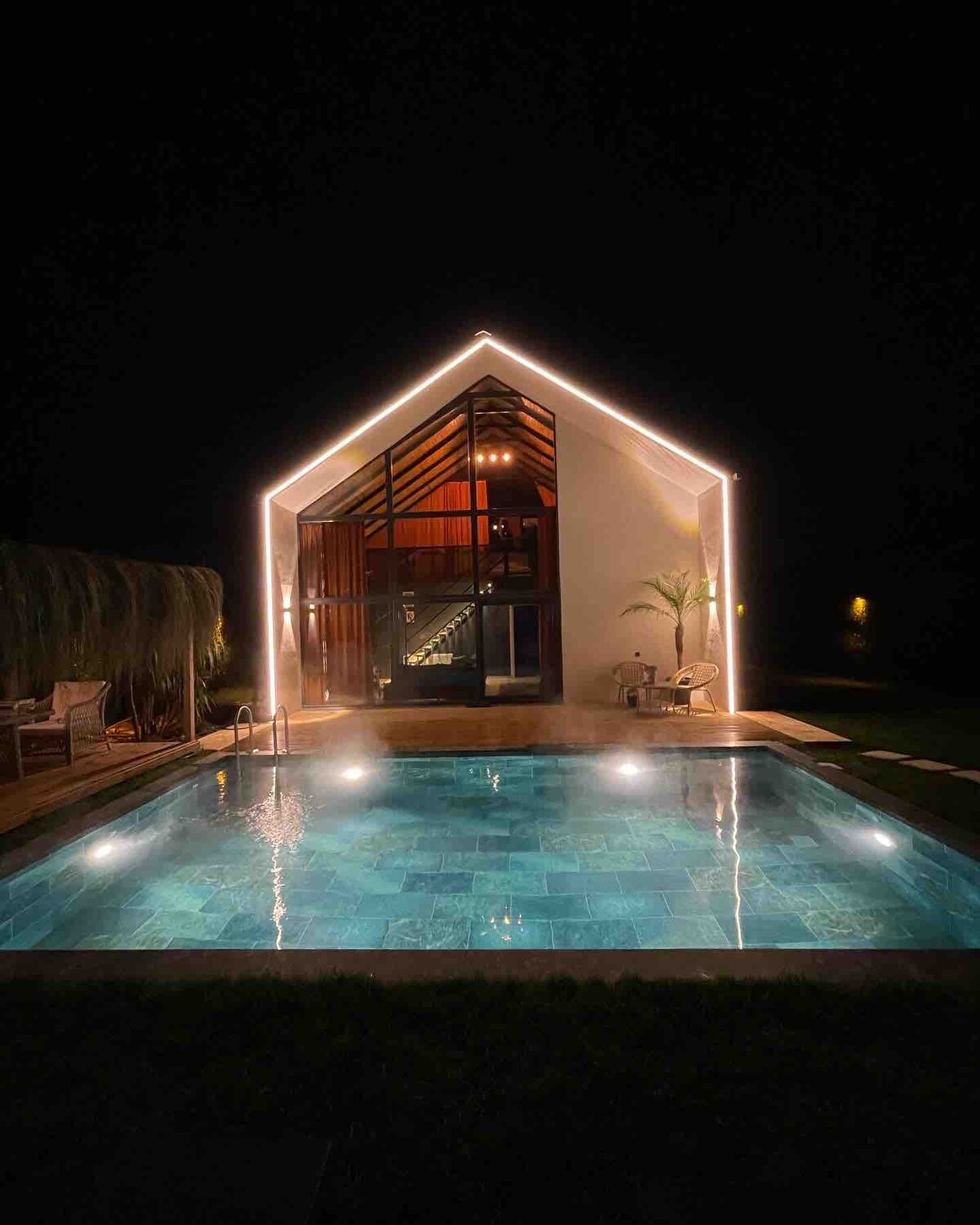 The modern villa is illuminated by soft lighting, highlighting its distinct architectural features. A sparkling pool reflects the charming glow of the lights, surrounded by a wooden deck. Lush greenery is visible in the background, enhancing the serene outdoor environment.