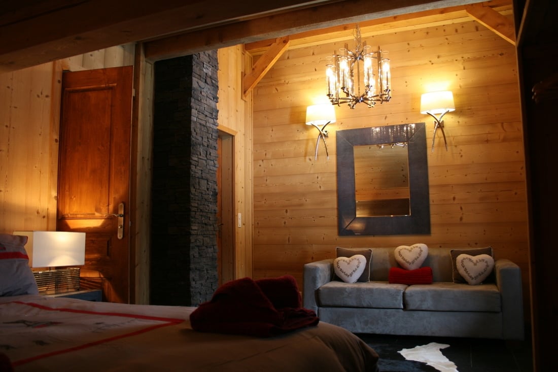 A warm wooden interior features a cozy sofa bed adorned with heart-shaped cushions. A stylish mirror above the sofa reflects soft lighting from wall sconces and a chandelier, enhancing the inviting ambiance. A plush blanket and bedside lamp complete the relaxing atmosphere.
