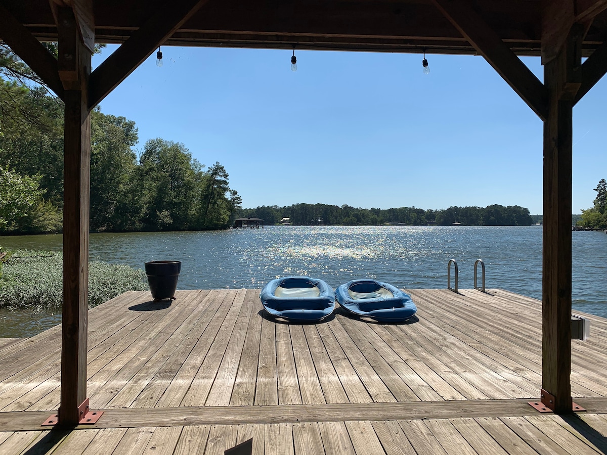 A wooden dock extends over the water, featuring two blue kayaks resting along the edge. Sunlight glimmers on the water's surface, surrounded by lush greenery. The serene environment invites leisure and exploration on Lake Sinclair.