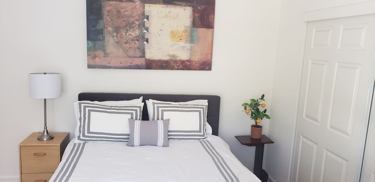 A serene bedroom is featured with a spacious bed adorned in white linens and decorative pillows. A modern artwork hangs above the bed, adding color to the room. A bedside lamp and a small plant stand with an orange tree enhance the inviting atmosphere.
