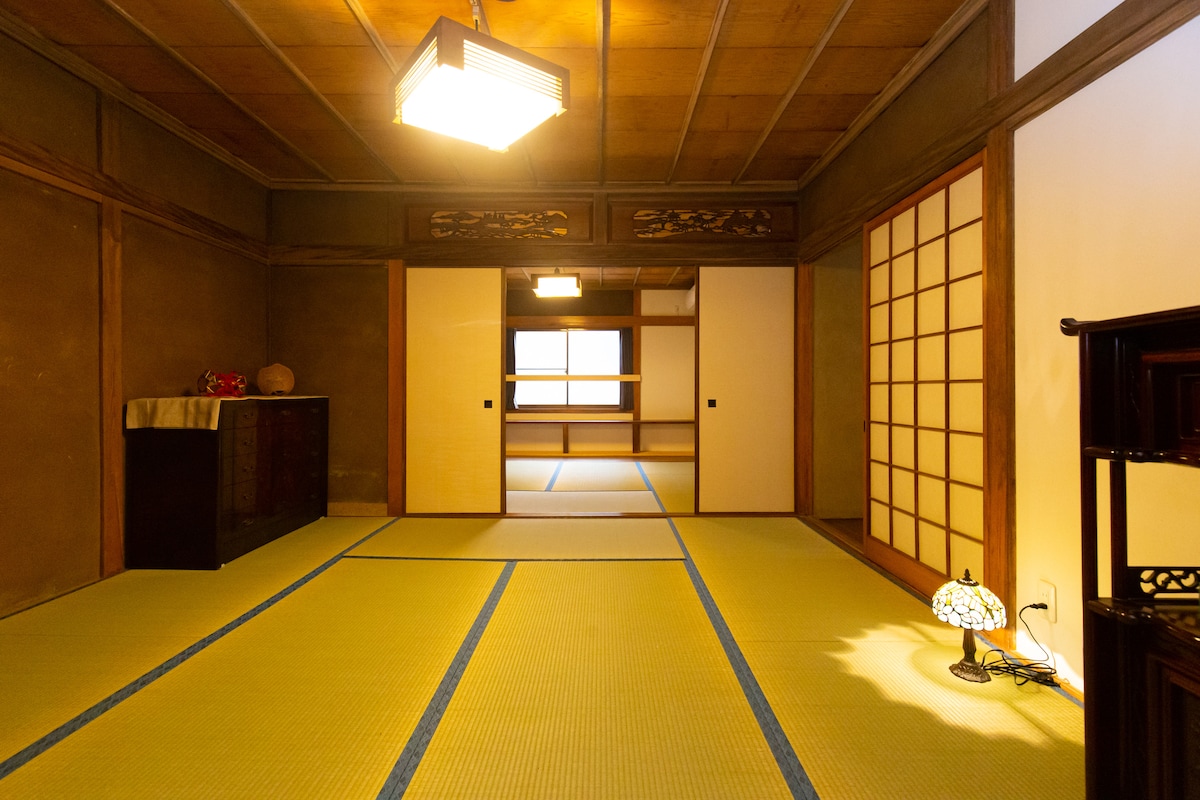 A spacious traditional Japanese room features tatami flooring and wooden accents. Soft lighting illuminates the space, highlighting shoji screens and a decorative lantern. A low table and a dark wooden chest are visible, enhancing the room's cultural ambiance.