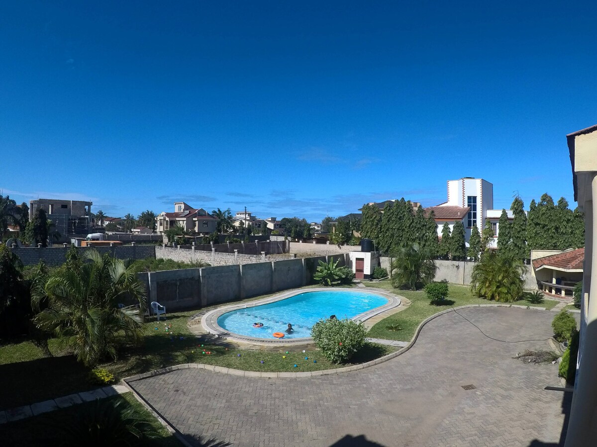 A clear view of a spacious pool surrounded by green landscaping is showcased. The area features palm trees and well-maintained gardens. The pool is bordered by a stone pathway, with a blue sky overhead and distant houses visible beyond the pool area.