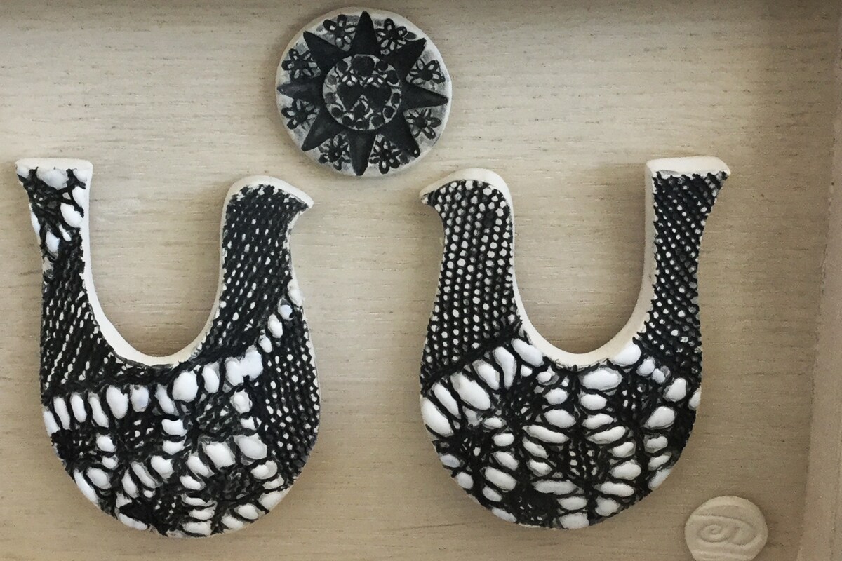 Two handmade decorative pieces are displayed against a light wooden background. The pieces, resembling stylized birds, are adorned with intricate black patterns and white detailing. Above them is a circular ornament featuring a star design, also in black and white, enhancing the artistic arrangement.