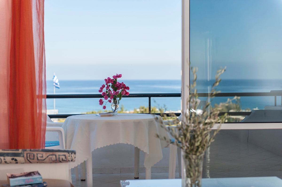A dining table adorned with a floral centerpiece is positioned near a large glass door, providing access to a private balcony. The view reveals the serene blue sea and the horizon. Soft orange curtains frame the window, allowing natural light to fill the space.