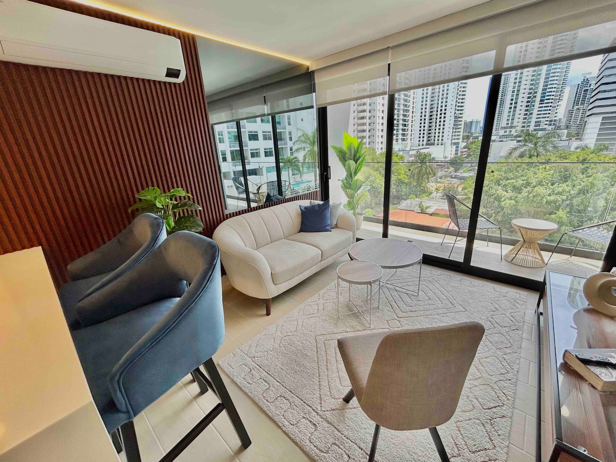 A modern living area features large glass doors that provide access to a balcony with city views. Comfortable seating includes a stylish sofa and two accent chairs. A round coffee table is positioned on a light area rug, complemented by a small plant for added greenery.