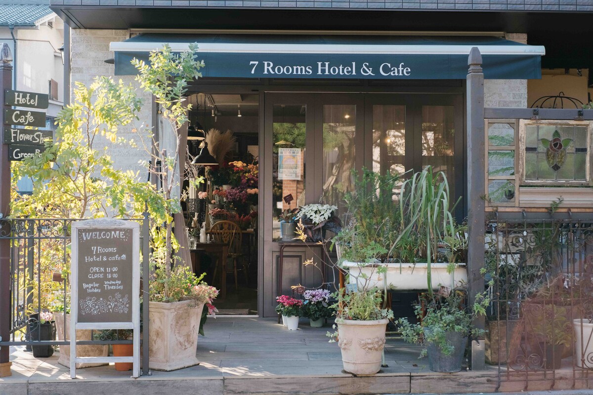 The entrance of 7 Rooms Hotel & Cafe features a welcoming canopy above large glass doors. Potted plants and flowers decorate the space, while a sign offers information about the hotel and cafe. The surrounding area is adorned with greenery, enhancing the inviting appearance.